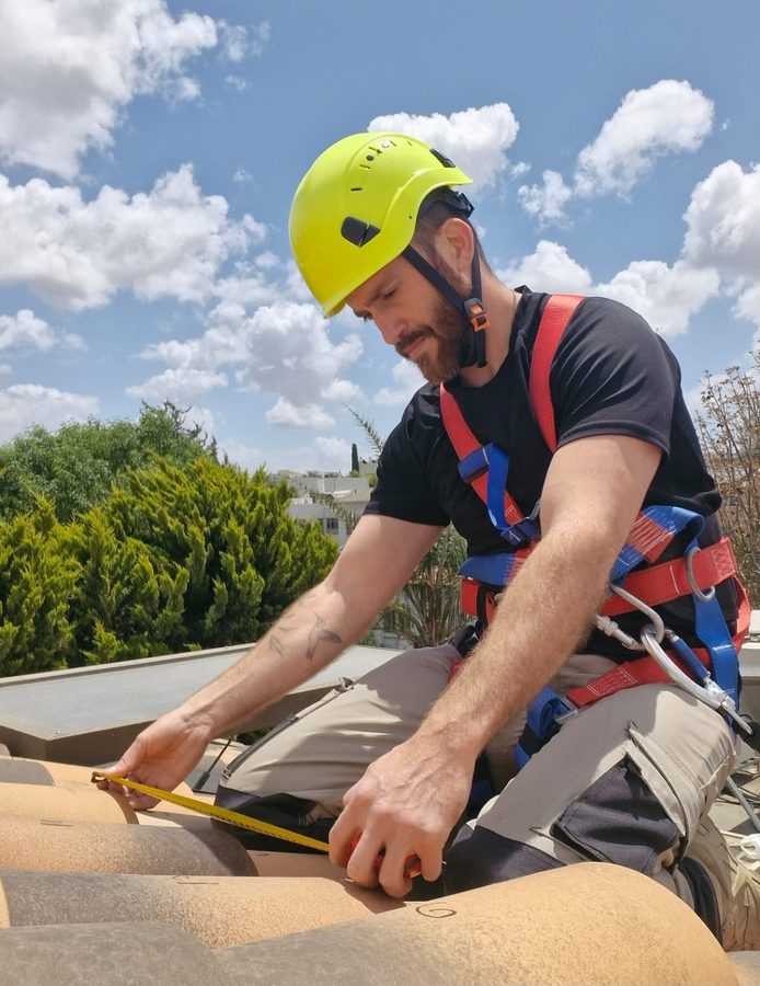 עידו בן גד יושב על גג רעפים בקסדה וברתמת בטיחות ומודד רעף בסרט מדידה